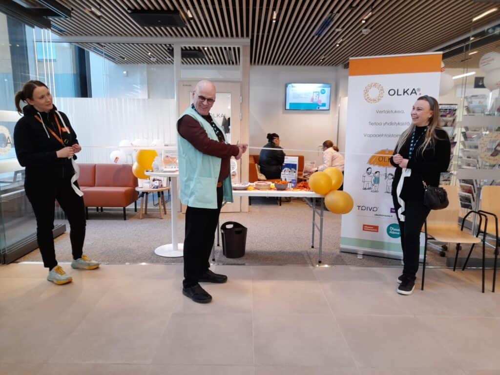 Kuva OLKA-pisteen avajaisista Assi-sairaalassa. Kuvassa keskellä vapaaehtoinen, joka leikkaa avajaisnauhaa. Sivuilla koordinaattorit pitelevät nauhasta kiinni. Takana Assi-sairaalan OLKA-piste, jossa avajaispäivän kunniaksi ilmapalloja. 